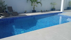 Piscina com azulejos azuis, cercada por espreguiçadeiras e plantas em vasos.