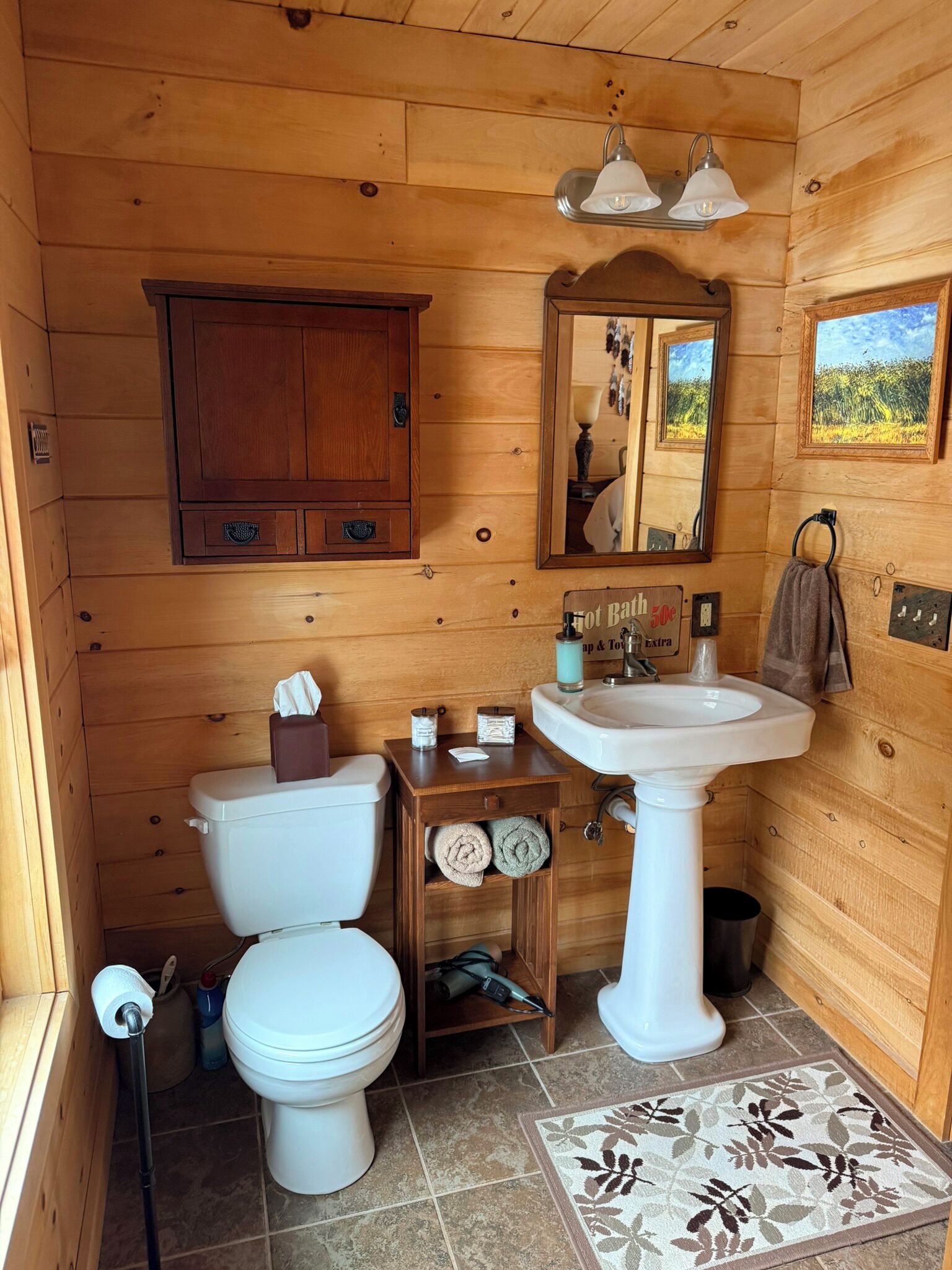 Une salle de bain rustique, aménagée dans une cabane en bois, comprenant des toilettes, un lavabo, un meuble en bois, un miroir et une petite étagère avec des serviettes.