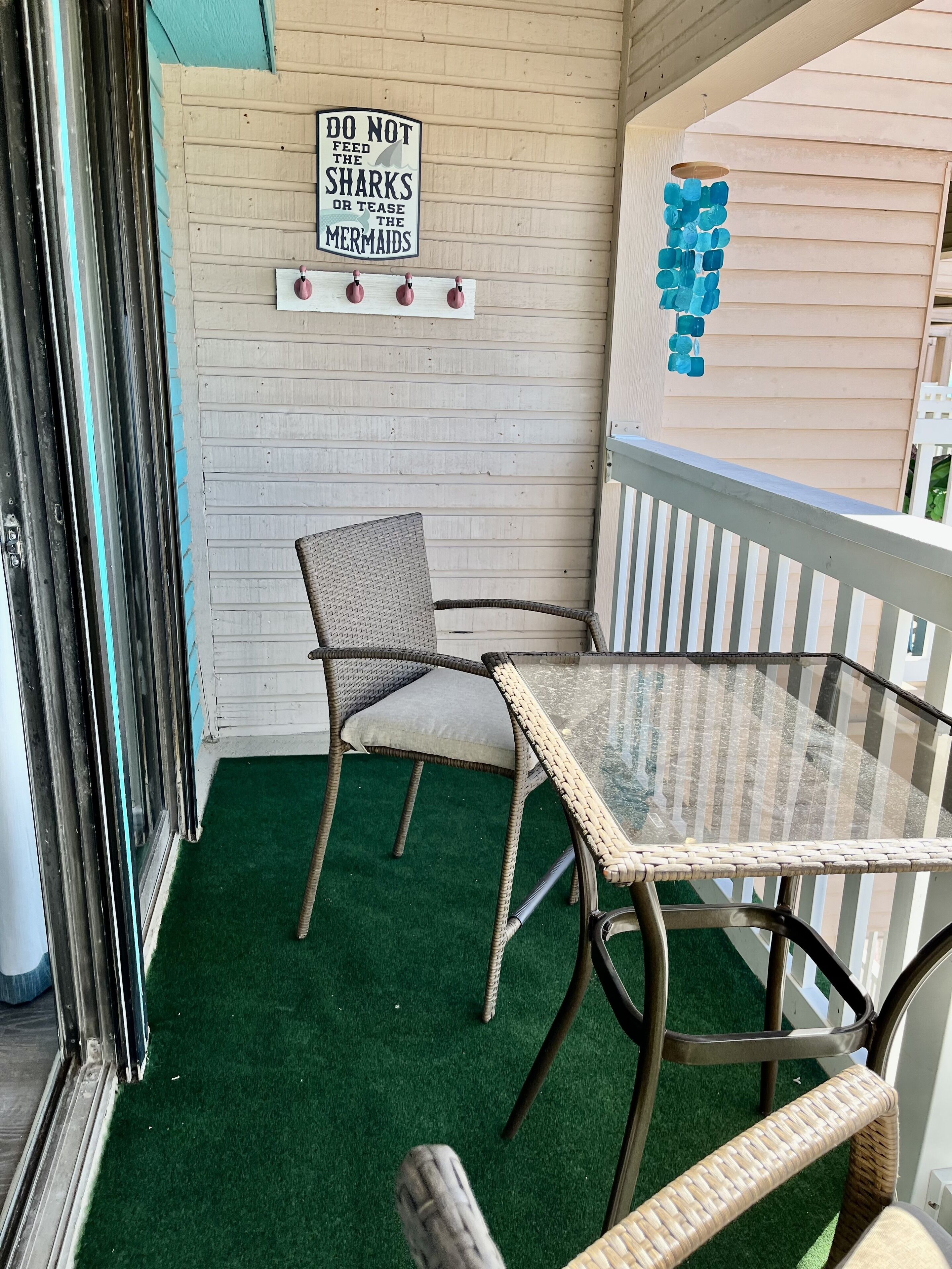 A corner of a room with a glass table and wicker chairs, a sign warning against feeding sharks or teasing mermaids, and a blue bottle decoration.