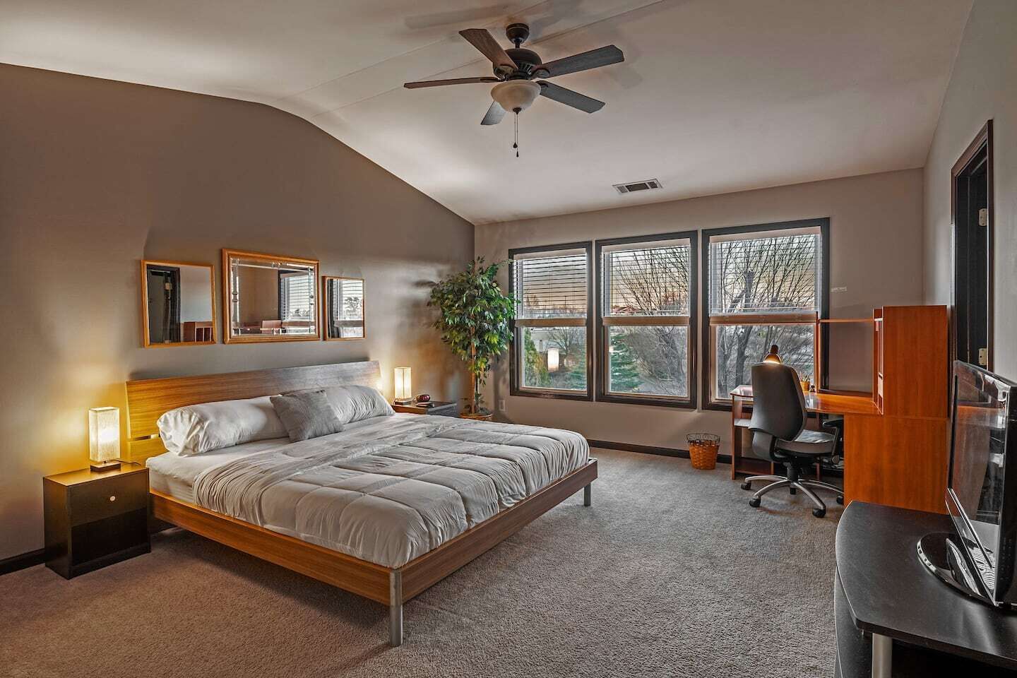 Une chambre à coucher comprenant un lit, des lampes de chevet, un bureau avec une chaise, une télévision et de grandes fenêtres.