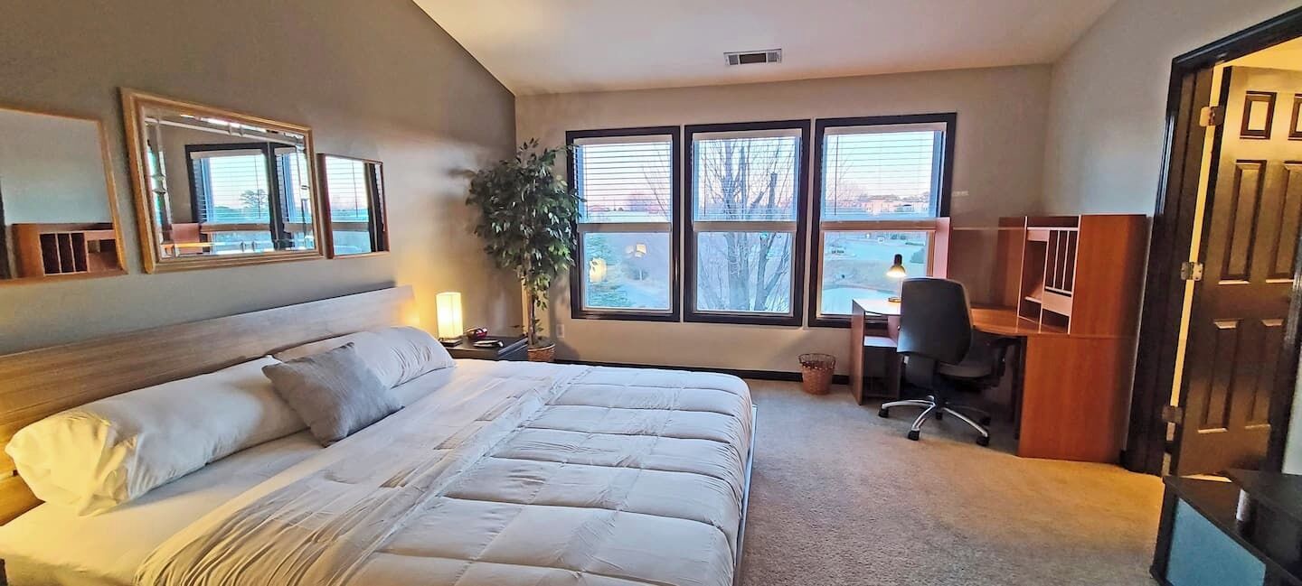 A bedroom with a bed, a desk, a chair, a potted plant, and large windows.