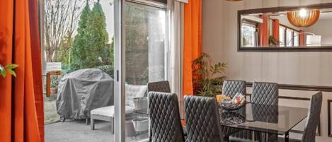 Un espace repas doté d’une porte vitrée donnant sur un coin salon extérieur abrité, une table à manger noire avec des chaises et un luminaire suspendu.