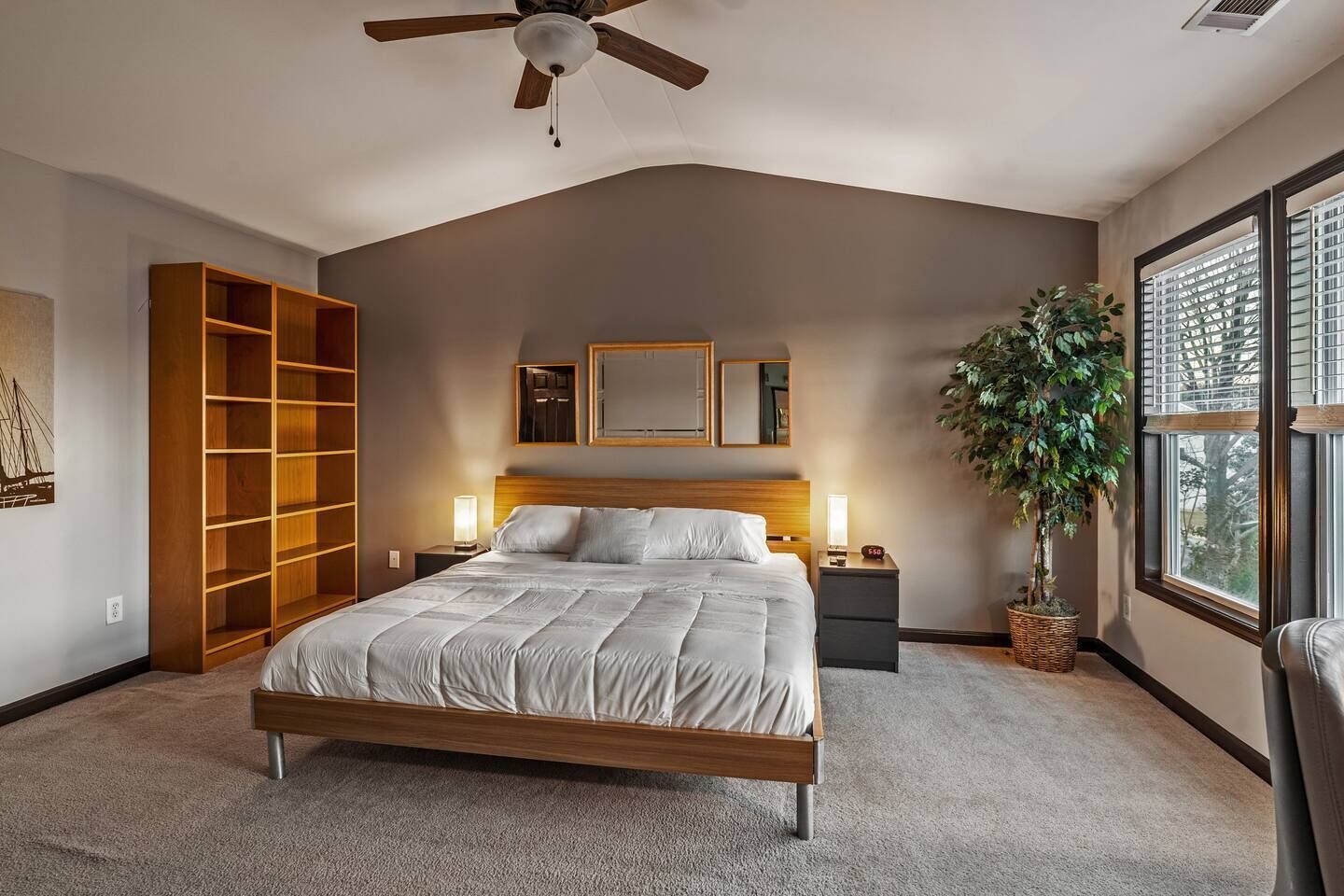 A bedroom with a bed, a ceiling fan, a nightstand, a potted plant, and a large window.