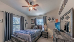 Une chambre à coucher avec un lit, un ventilateur de plafond, une commode, un téléviseur et une fenêtre avec des rideaux.