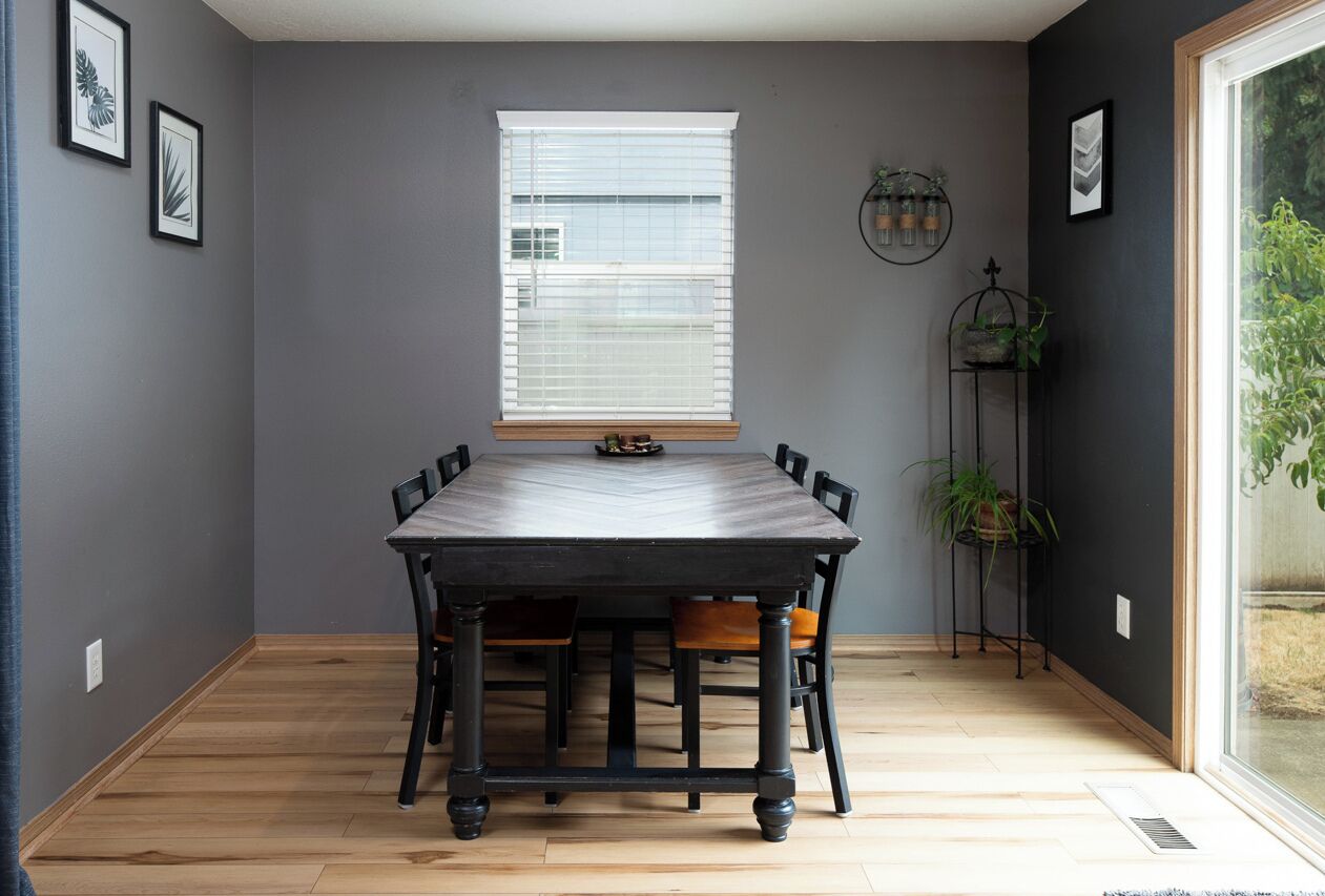 Une salle à manger avec une table et des chaises en bois foncé, une fenêtre avec des stores et une plante posée sur un support.