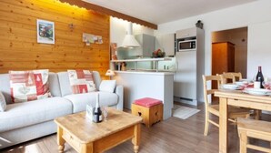 A cozy living room with a sofa, coffee table, and dining area with a table set for two. A kitchen with a microwave and cabinets is visible in the background. - Le Bec Rouge (Le Lac) by Interhome (Tignes)