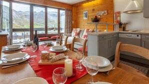 A dining area with a table set for four, a wooden wall, a kitchen area, and a view of mountains through large windows. - Le Bec Rouge (Le Lac) by Interhome (Tignes)