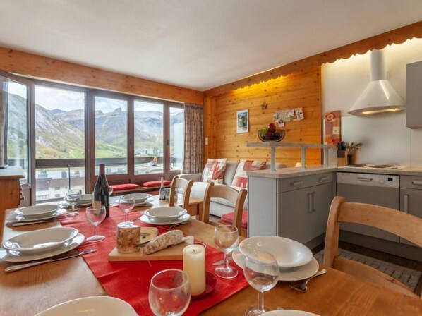 A dining area with a table set for four, a wooden wall, a kitchen area, and a view of mountains through large windows.