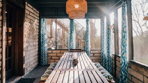 Un tavolo in legno con panche, un lampadario intrecciato a sospensione e tende a motivi decorativi in una stanza con pareti in mattoni e ampie finestre.