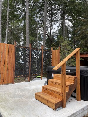 Une terrasse extérieure en bois, avec un escalier en bois menant à une zone abritée, le tout entouré d’une clôture en bois et d’arbres.
