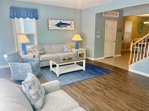 Uma sala de estar com sofá, poltrona, mesa de centro e luminárias. Há um quadro de um peixe na parede e uma placa com a inscrição 'BEACH HOUSE'.