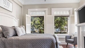 Une chambre avec un grand lit, deux fenêtres avec volets, une chaise et une cheminée.