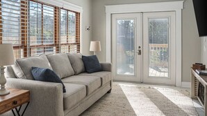 A living room with a beige sofa, two blue cushions, a wooden side table with a lamp, and a large window with blinds. - Old Dauphin Way Historic Home - Unit 6 (Mobile)