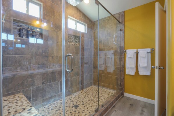A glass-enclosed shower with a tiled floor, a yellow wall, and a door with two towels hanging on a rack.