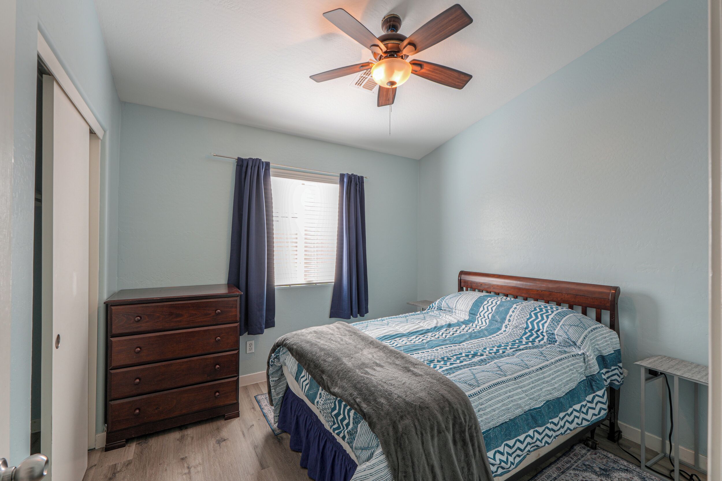 A bedroom with a bed, a dresser, a ceiling fan, and a window with curtains.