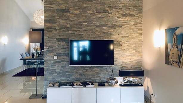 A modern living room with a stone wall, a flat-screen TV, a white entertainment unit, and a dining area with chairs and a table. - Stunning 3-Bed House in Tema-Ahenfie Villa (Tema Spintex)