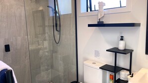 A modern bathroom with a glass shower enclosure, a toilet, a sink, and a small shelf with toiletries. - Gorgeous private studio apartment midway between Omaha & Matakana (Point Wells)