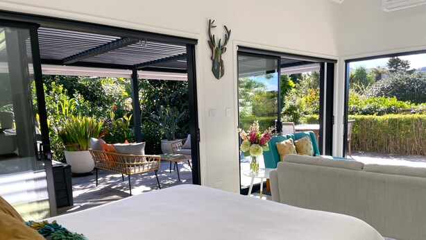 A bedroom with a large bed, a sofa, a dining table with chairs, and a view of a garden through open doors. - Gorgeous private studio apartment midway between Omaha & Matakana (Point Wells)