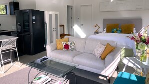 A modern room with a bed, a sofa, a glass coffee table, a kitchen area with a refrigerator, and a small table with a vase of flowers. - Gorgeous private studio apartment midway between Omaha & Matakana (Point Wells)