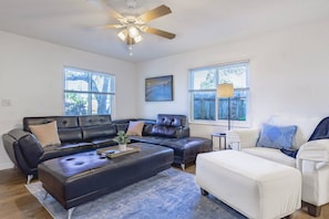 A modern living room with a sectional sofa, a white armchair, and a coffee table. There is a ceiling fan, large windows, and a framed picture on the wall. - Stunning Pool Home, Near Siesta Key & Dog Friendly (Sarasota)
