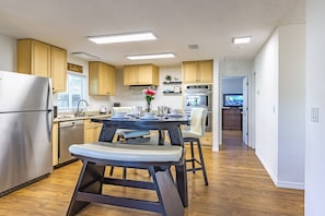 A modern kitchen with wooden cabinets, a stainless steel refrigerator, a dining table with chairs, and a television in the background.