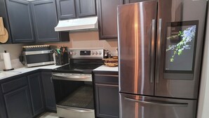 Una cocina moderna con armarios oscuros, electrodomésticos de acero inoxidable, incluyendo un refrigerador, horno y microondas, y una pantalla digital en el refrigerador.
