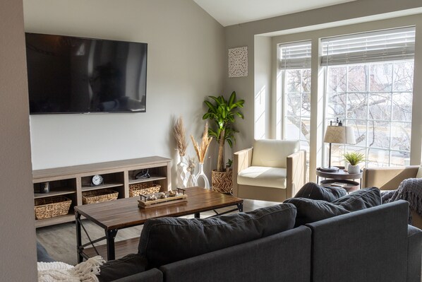Una sala de estar moderna con una televisión de pantalla plana, una mesa de centro de madera, un sofá y una silla. Hay plantas decorativas y una lámpara sobre una mesita auxiliar.