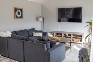 Una sala de estar moderna con un sofá seccional gris, un televisor de pantalla plana, una mesa consola de madera y una lámpara de pie.