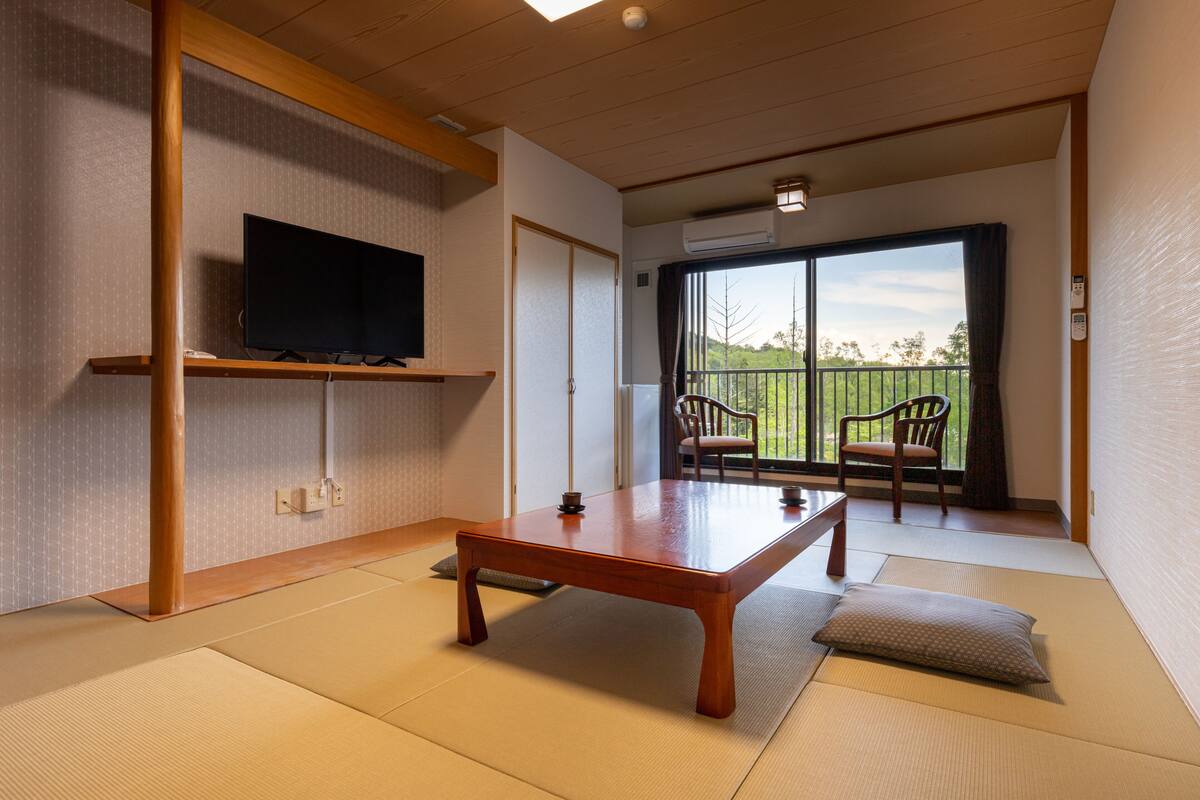 Una habitación japonesa tradicional con suelo de tatami, una mesa de madera baja, un televisor en un estante y un balcón con sillas.