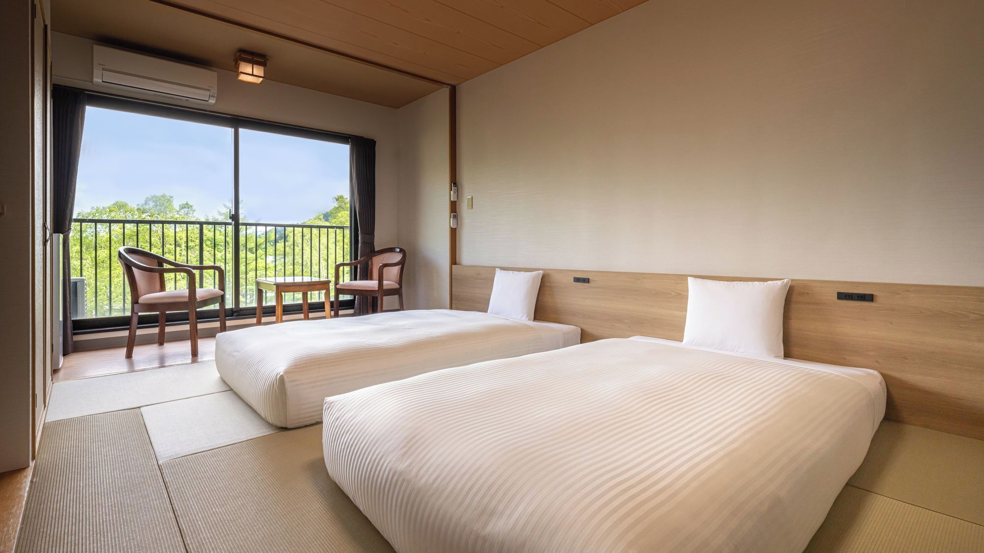 A hotel room with two beds, a balcony with chairs, and a view of greenery.