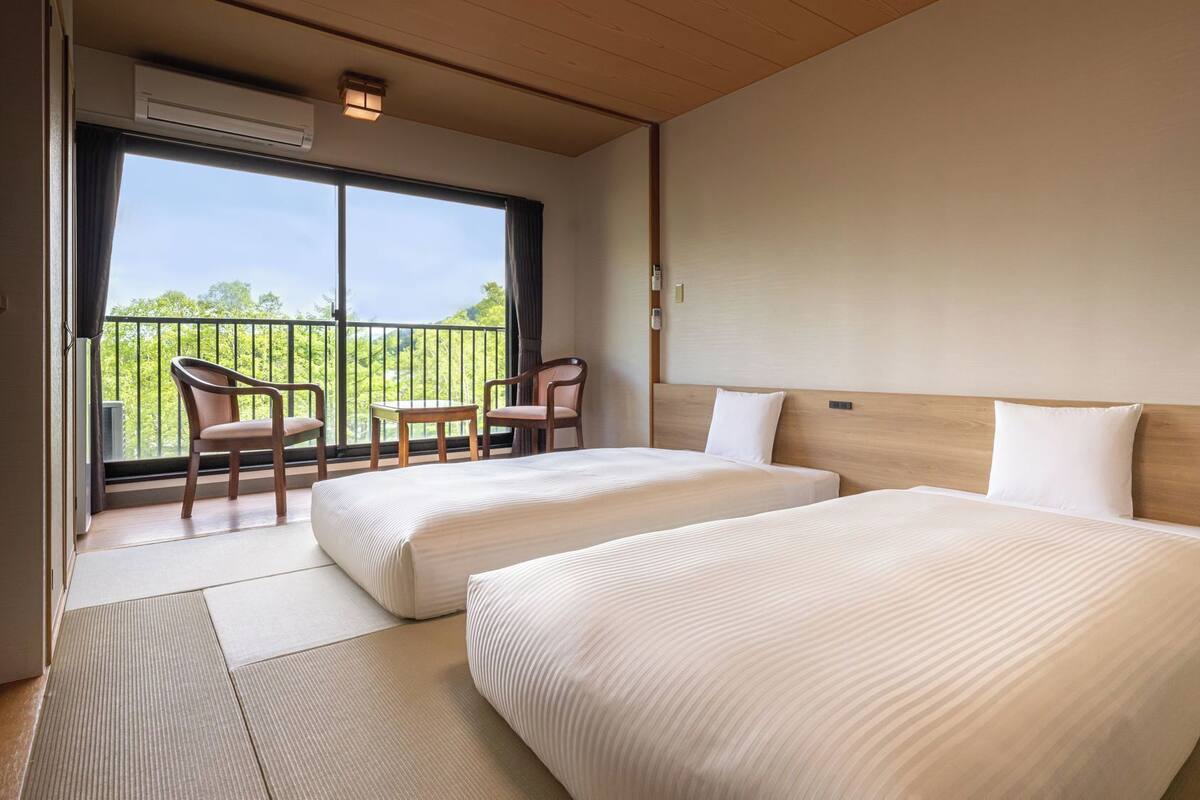 Habitación de hotel con dos camas, un balcón con sillas y vistas a zonas verdes.