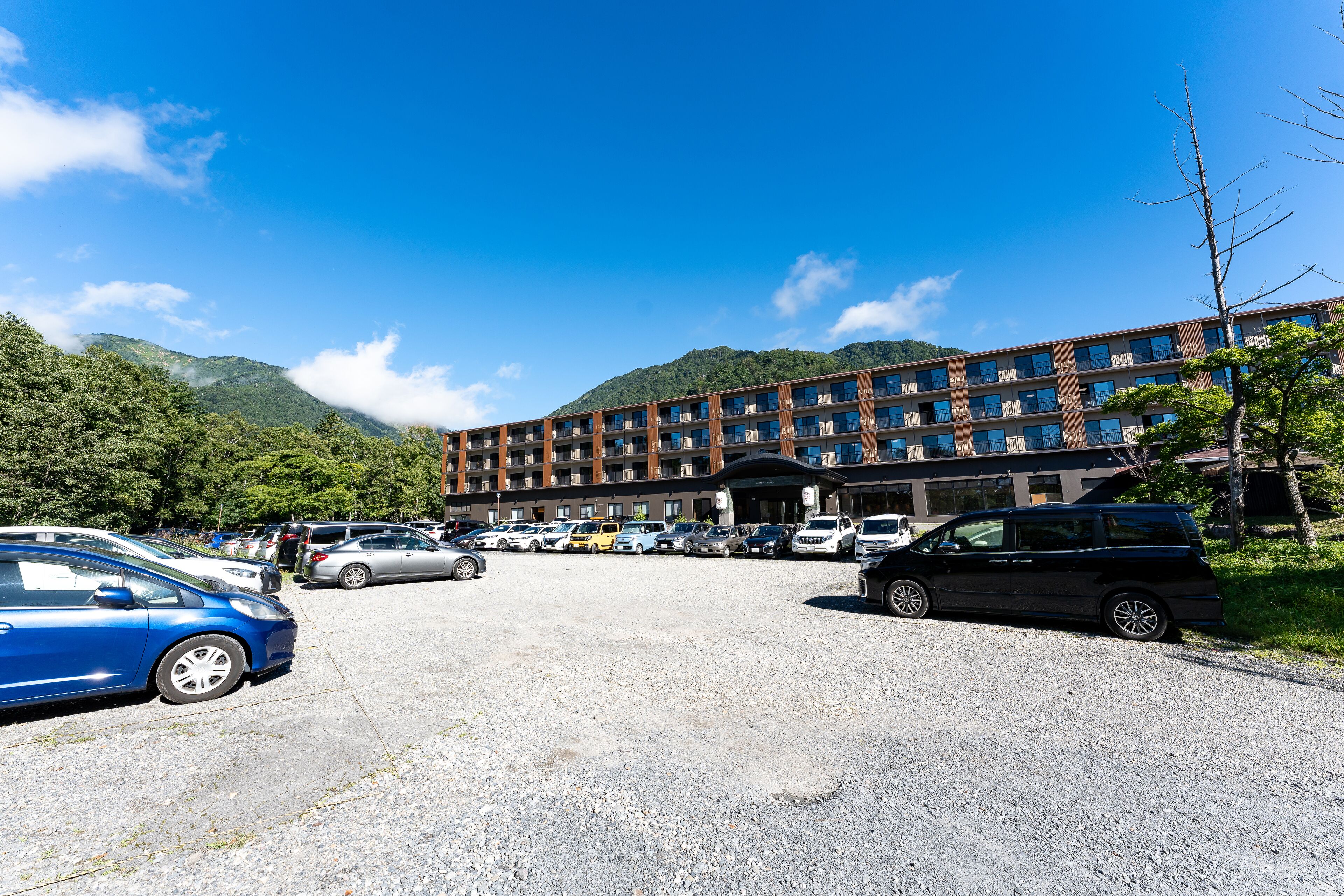 A multi-story hotel with a parking lot in front, surrounded by mountains and greenery.