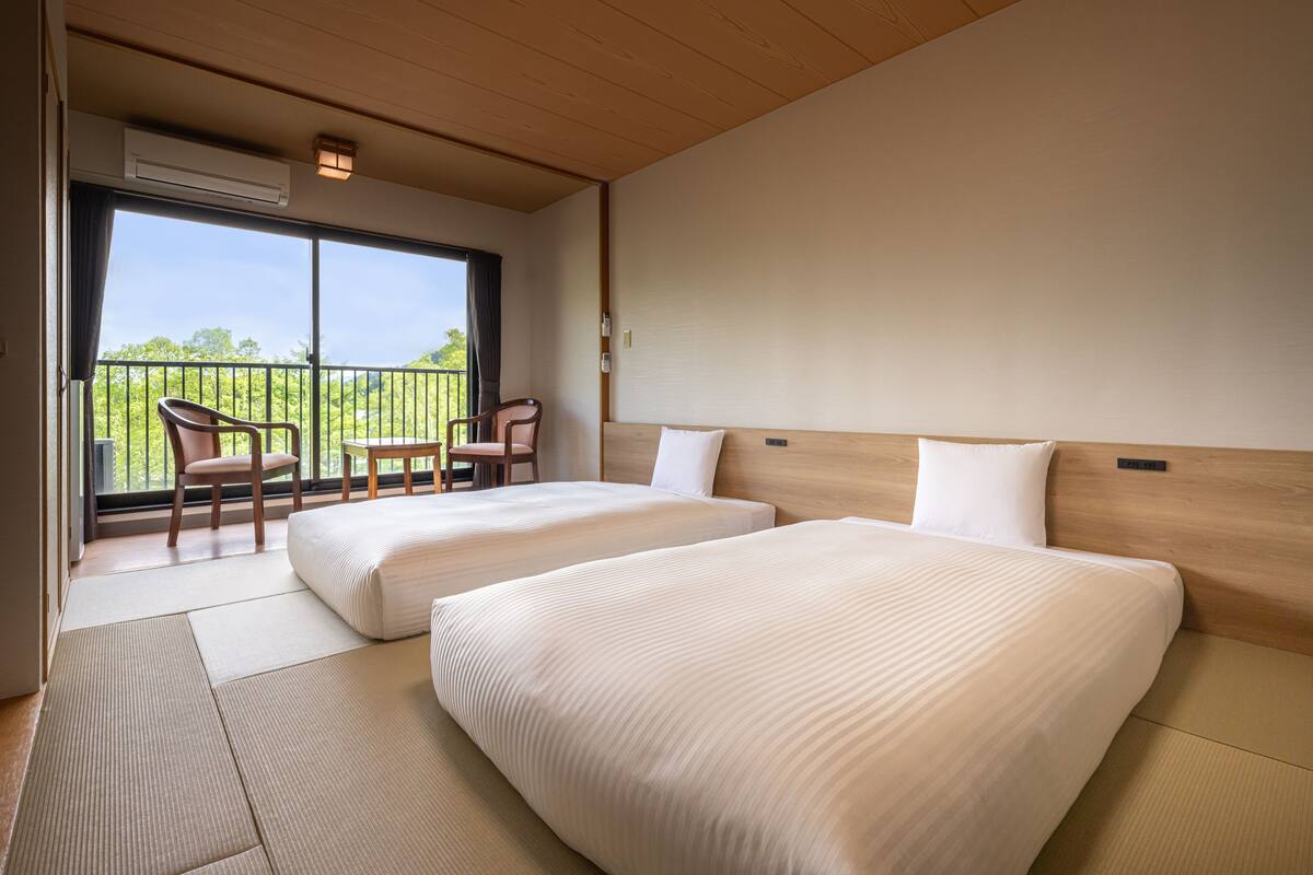 Habitación de hotel con dos camas, un balcón con sillas y vistas a zonas verdes.