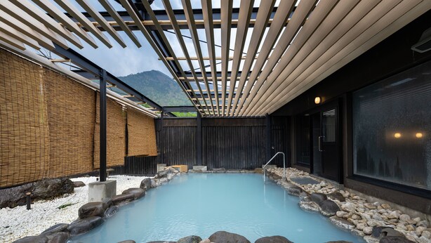 A traditional Japanese onsen with a wooden deck, a stone bath surrounded by rocks, and a view of mountains in the background.