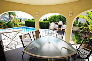 Un patio soleado con una mesa de vidrio, sillas y vistas a una piscina y follaje.