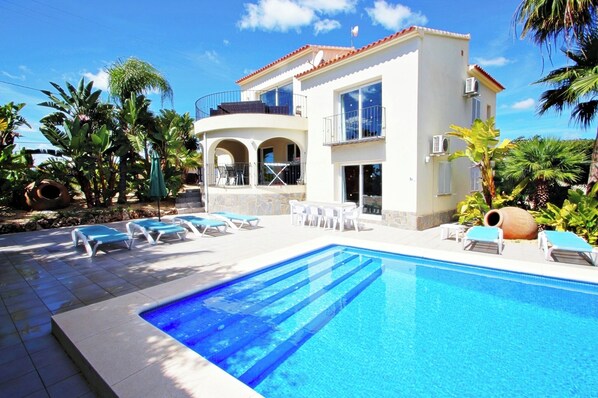 A two-story house with a swimming pool, outdoor furniture, and tropical plants.