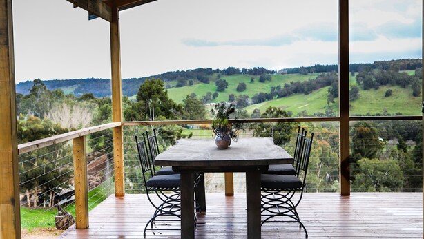 Une terrasse en bois avec une table et des chaises, donnant sur une vallée verdoyante parsemée d’arbres et de collines.
