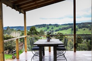 A wooden deck with a table and chairs, overlooking a green valley with trees and hills.