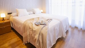 Una cama doble con una colcha blanca y dos almohadas, una mesita de noche con una lámpara, y una mesita auxiliar de madera.