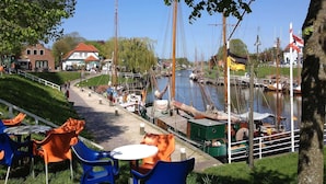 Un café situé au bord de la rivière, avec une terrasse, un quai où sont amarrés des bateaux et un bâtiment surmonté d’un toit rouge.