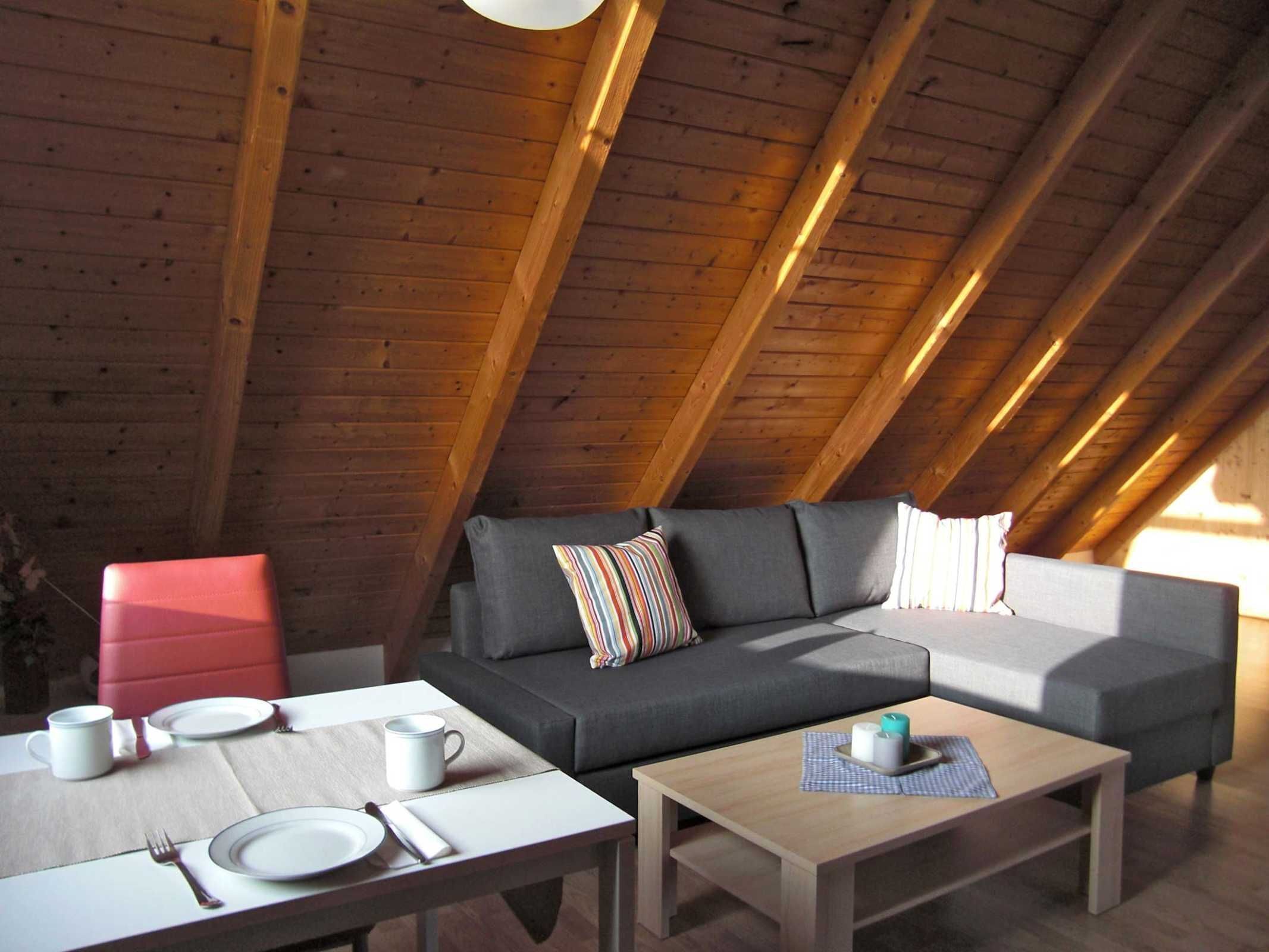 Een zolderkamer met een houten plafond, een grijze bank, een rode stoel, een klein tafeltje met een witte mok en een bijzettafeltje met kaarsen.