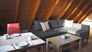 Une pièce mansardée avec un plafond en bois, un canapé gris, un fauteuil rouge, une petite table avec une tasse blanche et une table basse ornée de bougies.
