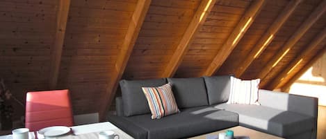 An attic room with a wooden ceiling, a grey sofa, a red chair, a small table with a white mug, and a coffee table with candles.