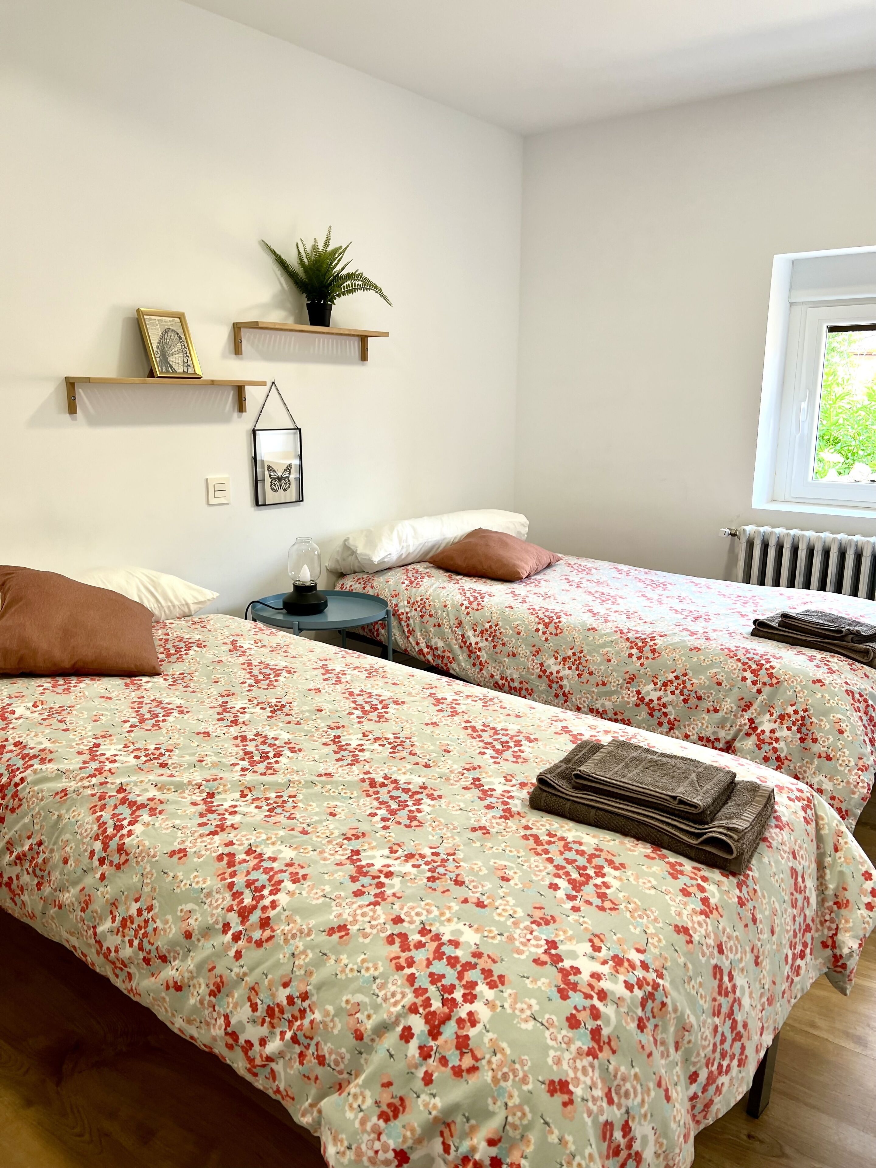 Ein Schlafzimmer mit zwei Betten, jeweils mit floraler Bettwäsche, einem kleinen Tisch und einem Stuhl. An der Wand befinden sich Regale mit Dekoration und ein Fenster mit Blick auf Grünflächen.