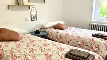 Ein Schlafzimmer mit zwei Betten, jeweils mit floraler Bettwäsche, einem kleinen Tisch und einem Stuhl. An der Wand befinden sich Regale mit Dekoration und ein Fenster mit Blick auf Grünflächen.