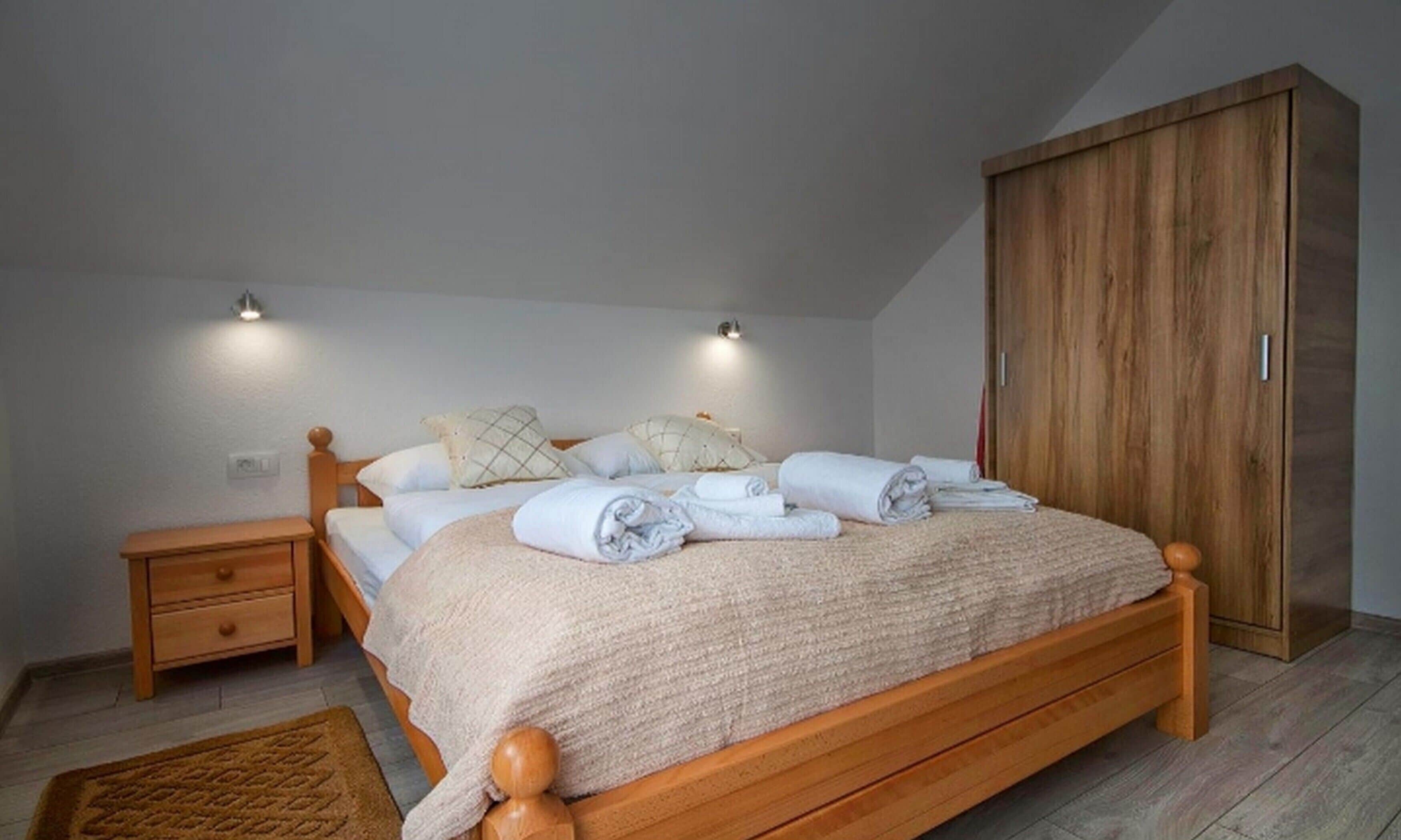A neatly made bed with rolled towels, a wooden nightstand, a wardrobe, and a small rug.