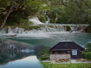 Un chalet doté de fondations en pierre et d’un toit sombre, situé au bord d’un lac aux eaux claires, avec une cascade en arrière-plan.