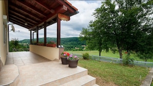 A covered outdoor area with steps leading to a terrace, potted plants, and a view of a green landscape with trees and hills. - Apartment Josipa (Rakovica)