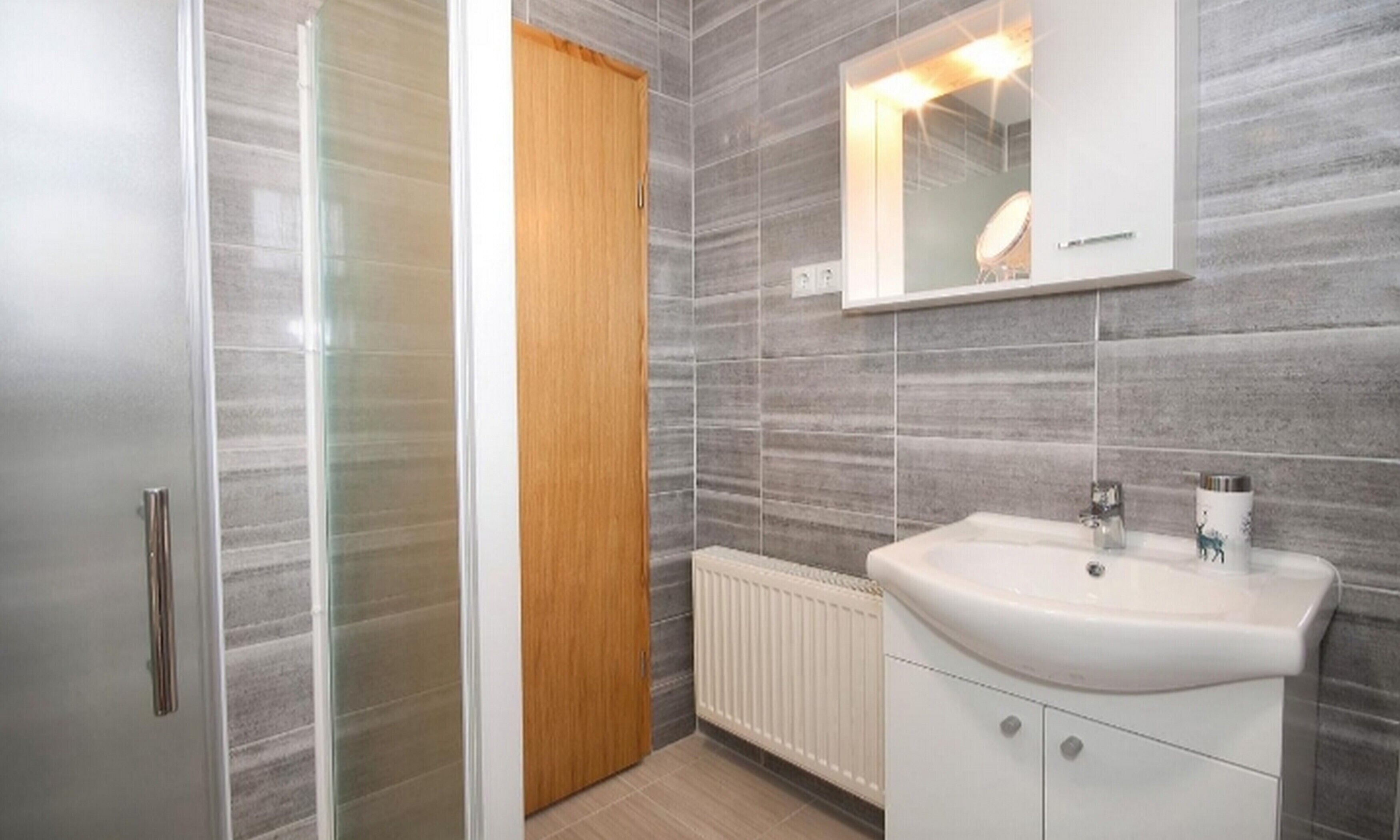 A bathroom with a glass shower enclosure, a wooden door, a white sink with a mirror above it, and grey tiled walls.