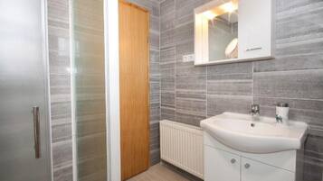 A bathroom with a glass shower enclosure, a wooden door, a white sink with a mirror above it, and grey tiled walls.
